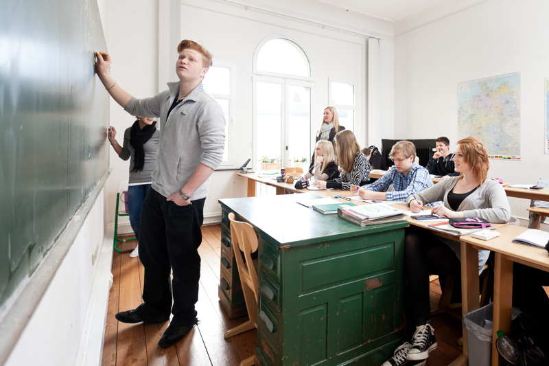 Ein Schüler steht vor einer kleinen Klasse an der Tafel und denkt über die Aufgabenstellung nach.